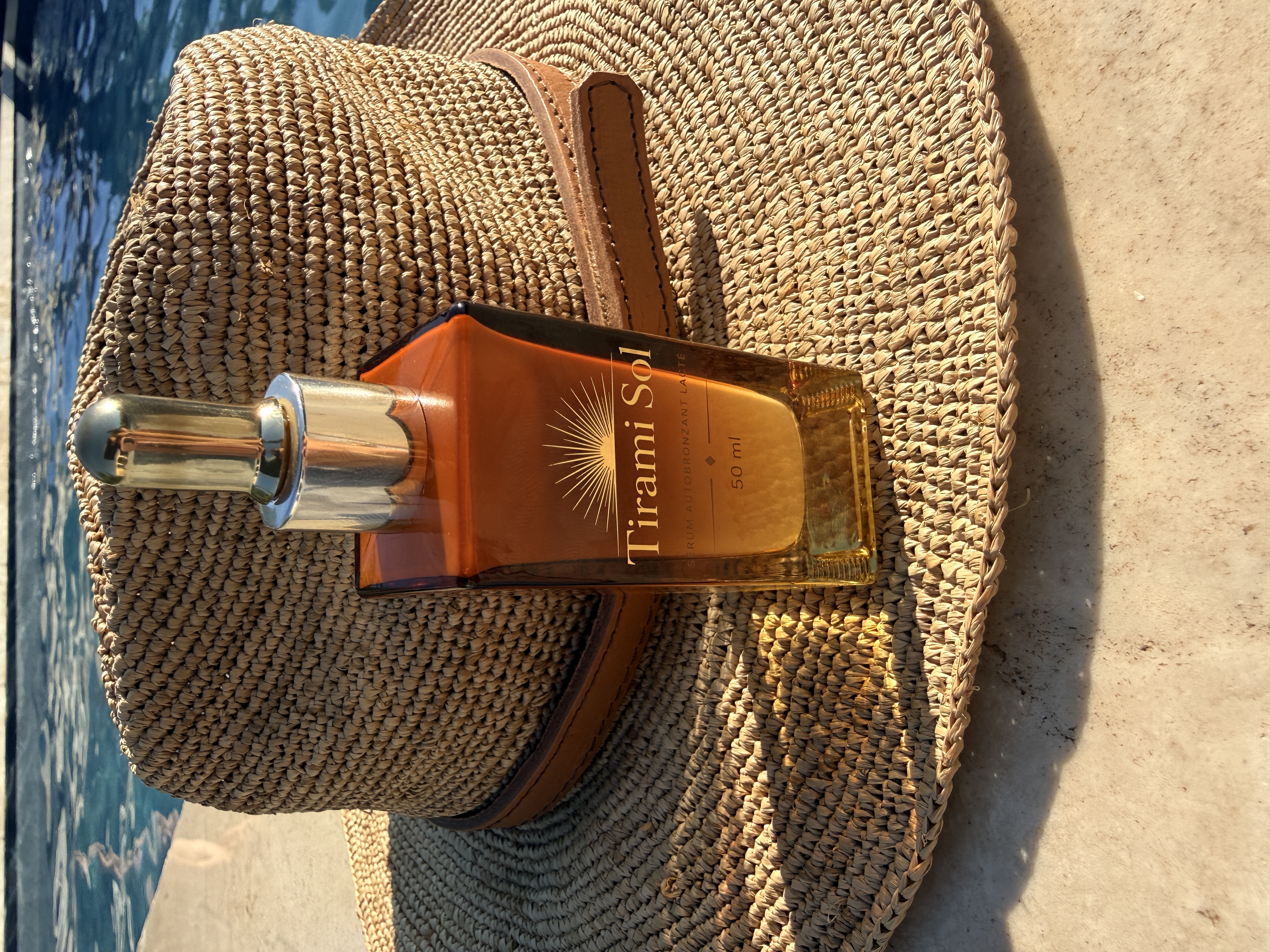 Photo du véritable flacon de sérum autobronzant TiraMiSol 50 ml et de sa pipette dorée, devant un chapeau
